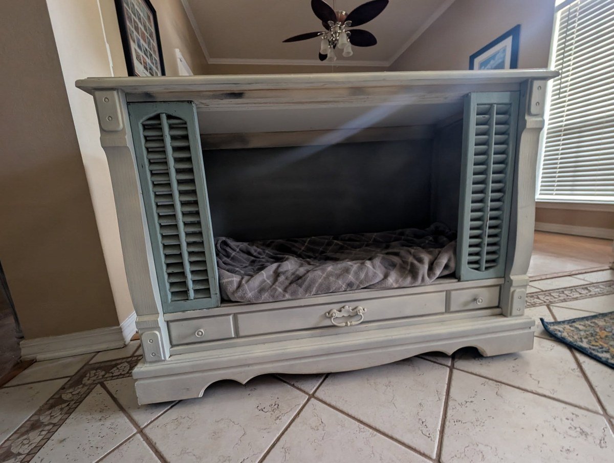 DIY pet bed made from an old wooden console TV cabinet, painted gray with blue shutter doors and a blanket inside. No readable text.