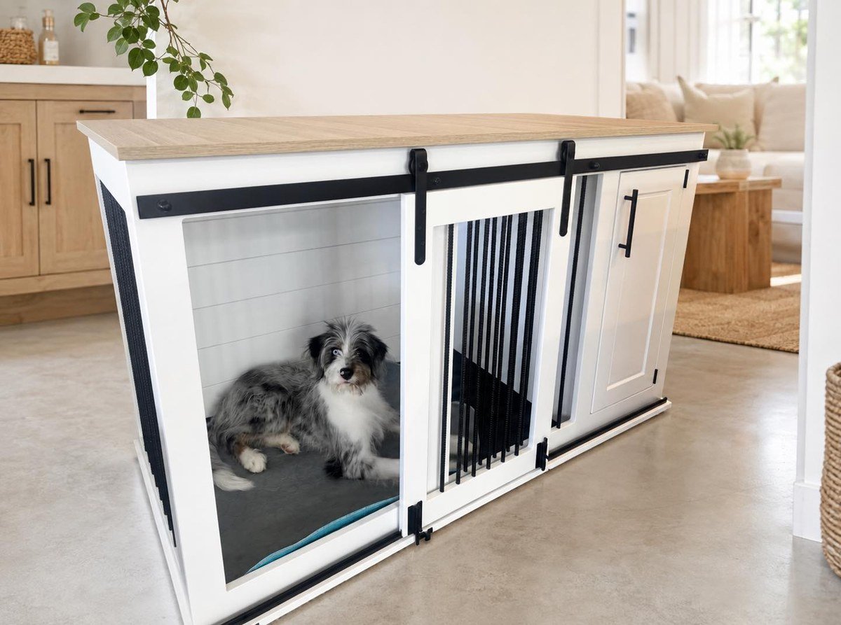 White kennel with natural timber top and matte black sliding barn-door hardware, fluffy merle dog lying inside. No readable text per visual summary.