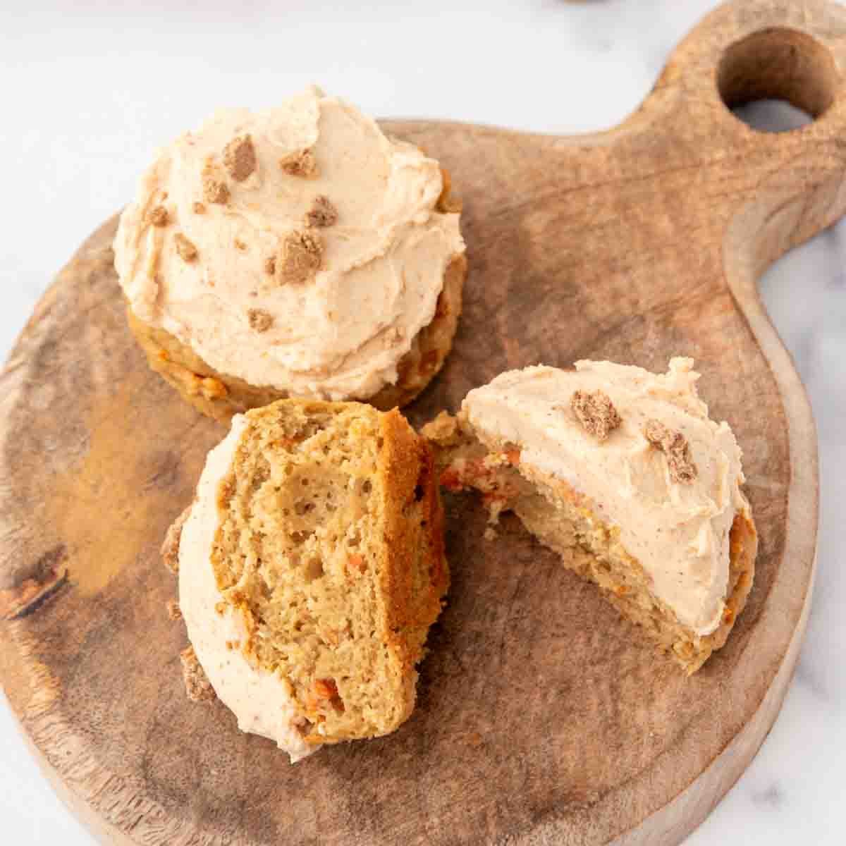 Frosted carrot pupcakes with crushed biscuit topping on a wooden board, one cut open showing the crumb