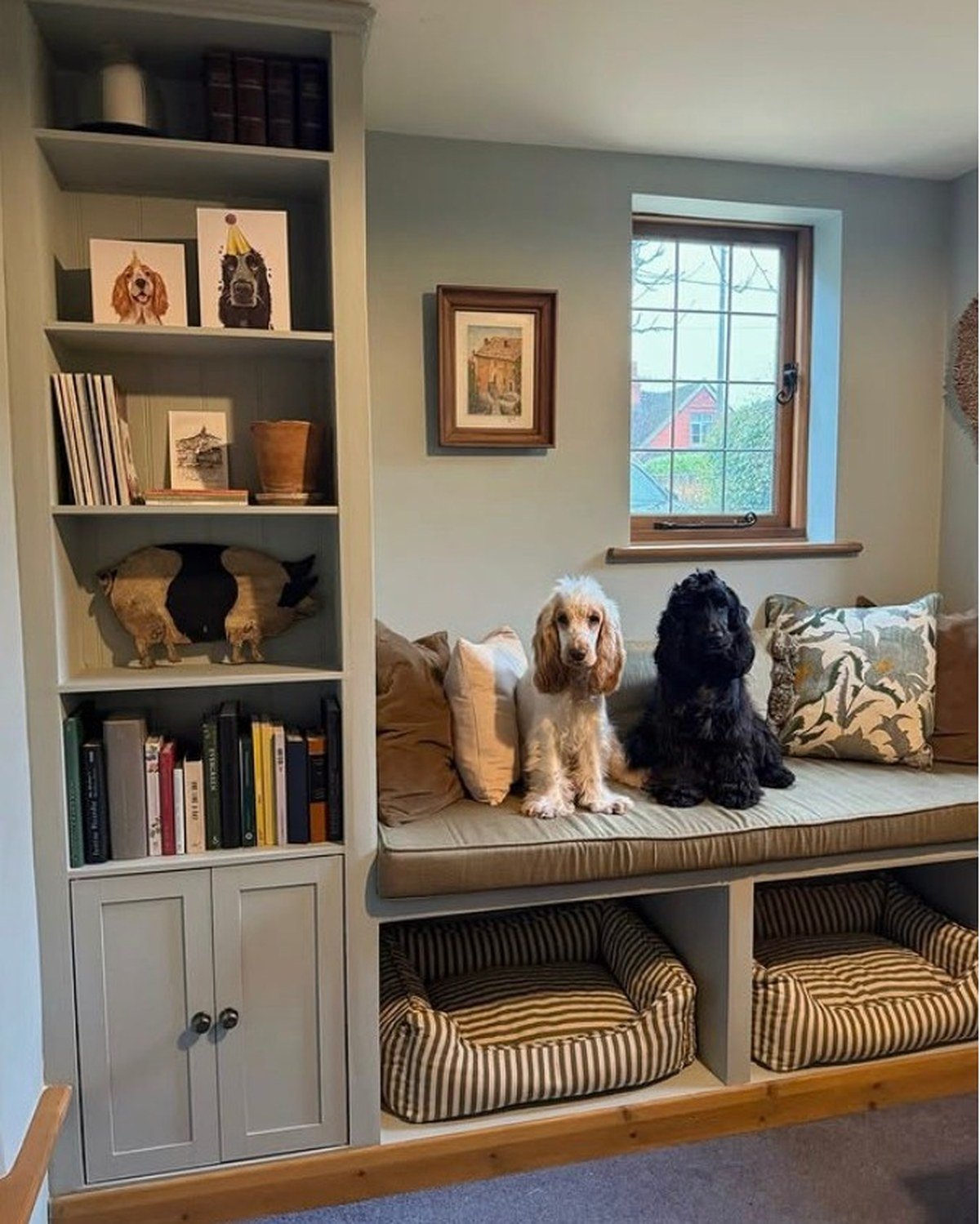 Custom reading nook with cushioned window seat, two cocker spaniels on pillows, bookshelves and dog beds in cubbies below. No readable text per visual summary.
