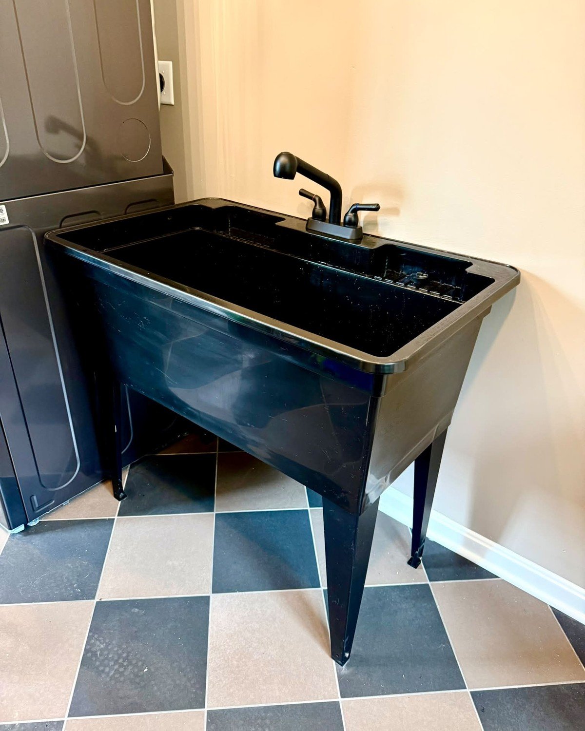 Newly installed black freestanding laundry utility tub with a matte black faucet in a laundry room corner; author caption notes it will be used for dog showers. No text visible in the image. (No dog in frame.)