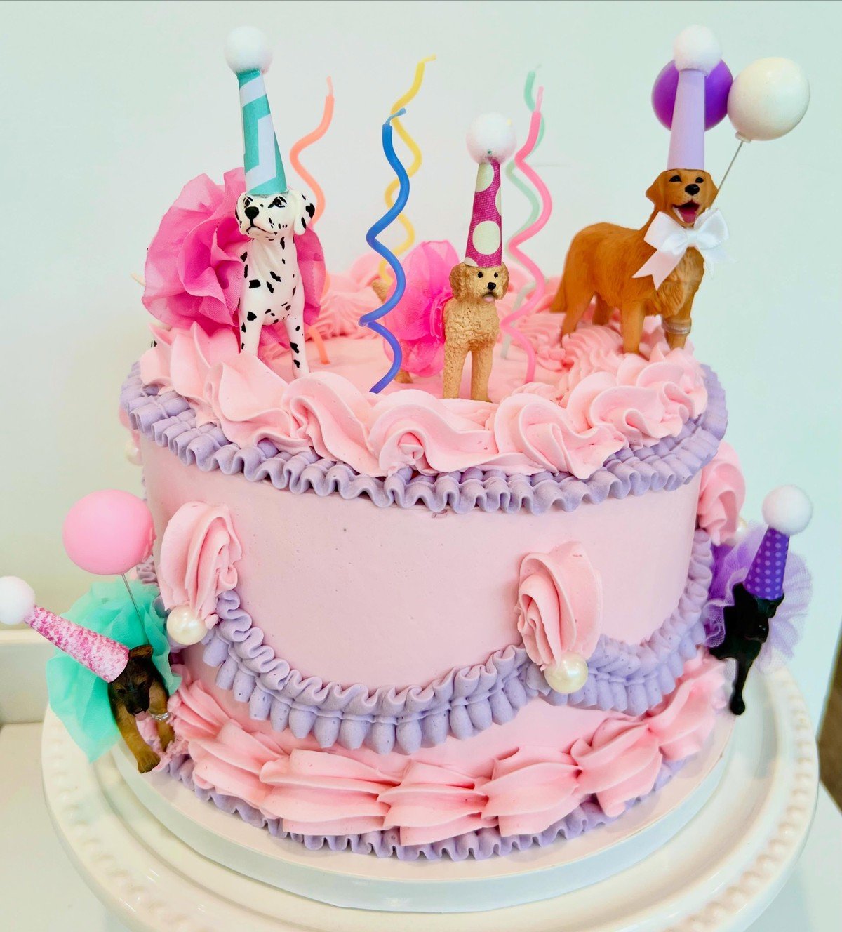 A two-tier pink and lavender cake topped with five toy dog figurines wearing party hats and tutus.