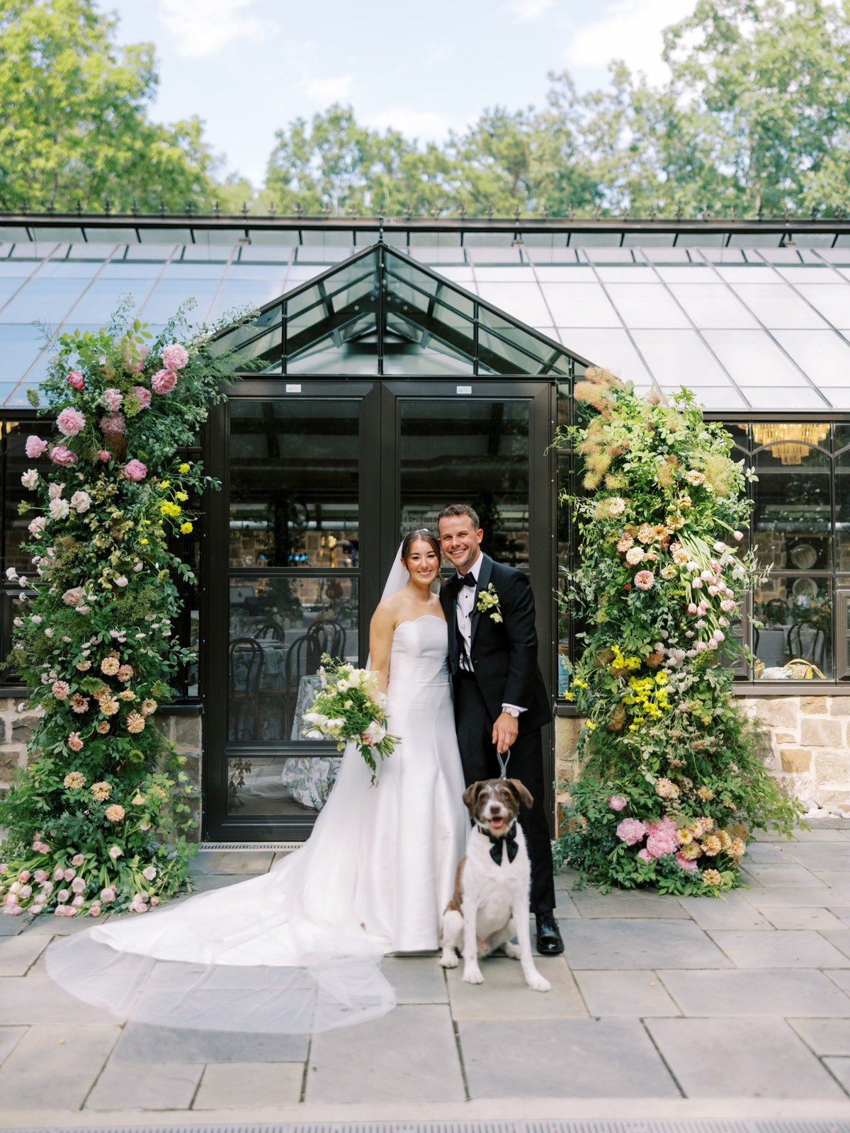 Bride and groom posing with their dog in a bow tie in front of a flower-adorned greenhouse