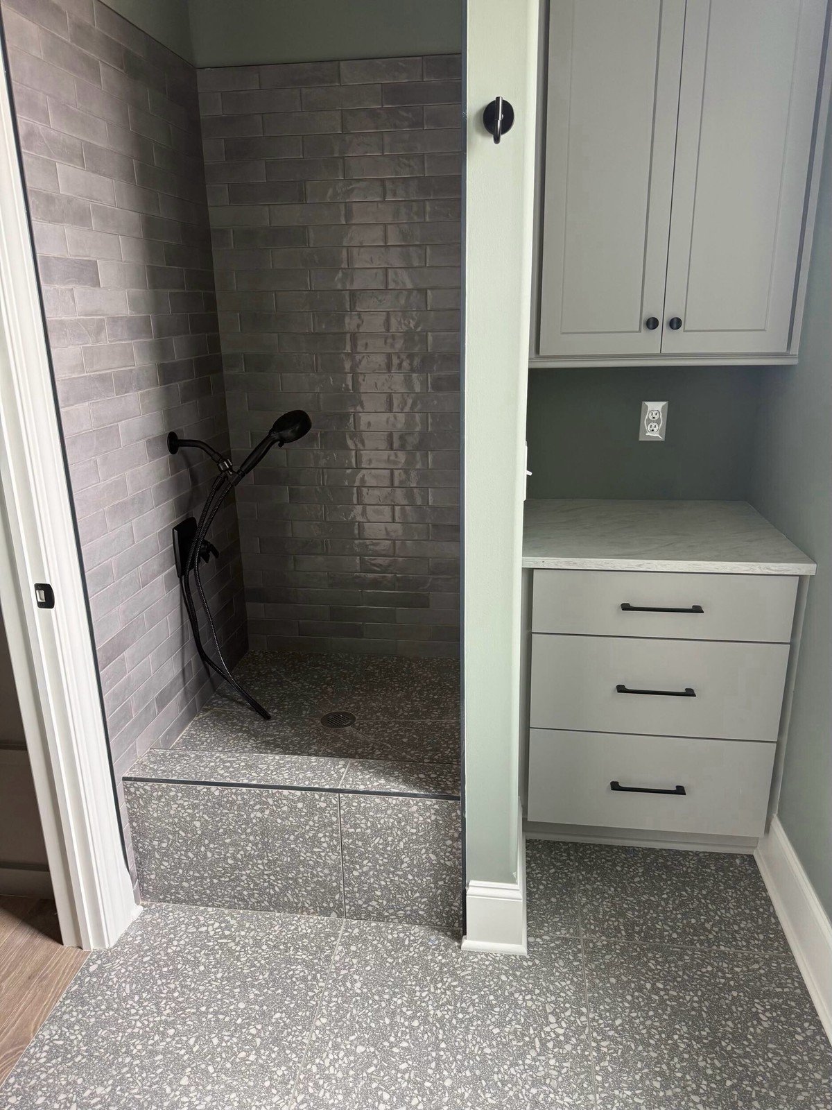 Mudroom corner with a raised gray-subway-tile shower area, speckled terrazzo floor and matte black wall-mounted handheld showerhead beside storage cabinetry - a hardworking wet corner. No text visible in the image.