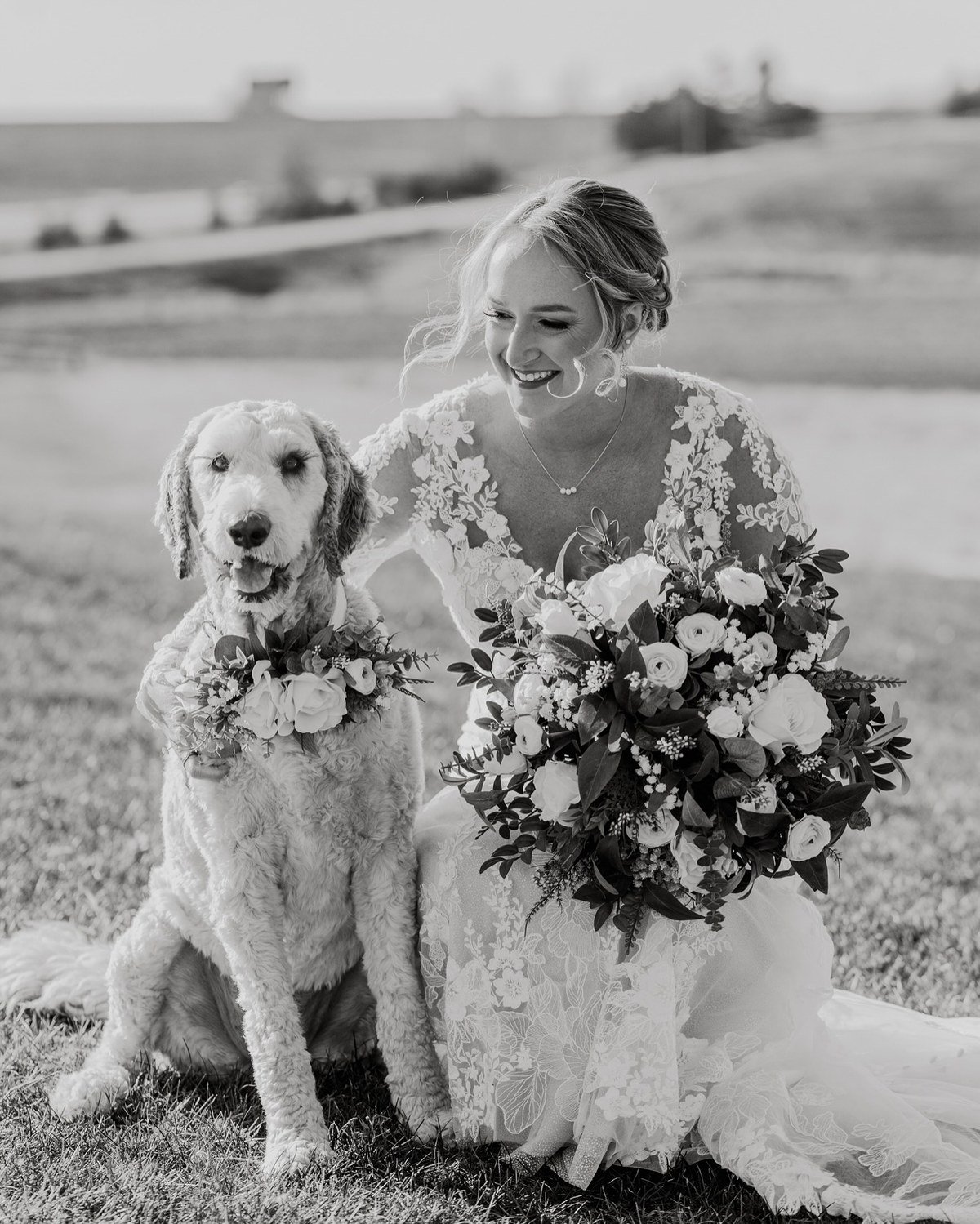 Bride smiling beside her white dog wearing a flower collar in a black-and-white wedding portrait