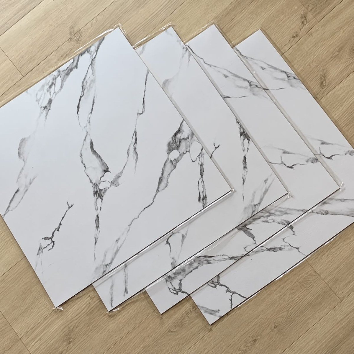 Four marble-pattern peel-and-stick vinyl floor tiles overlapping on wood laminate flooring. No readable text.