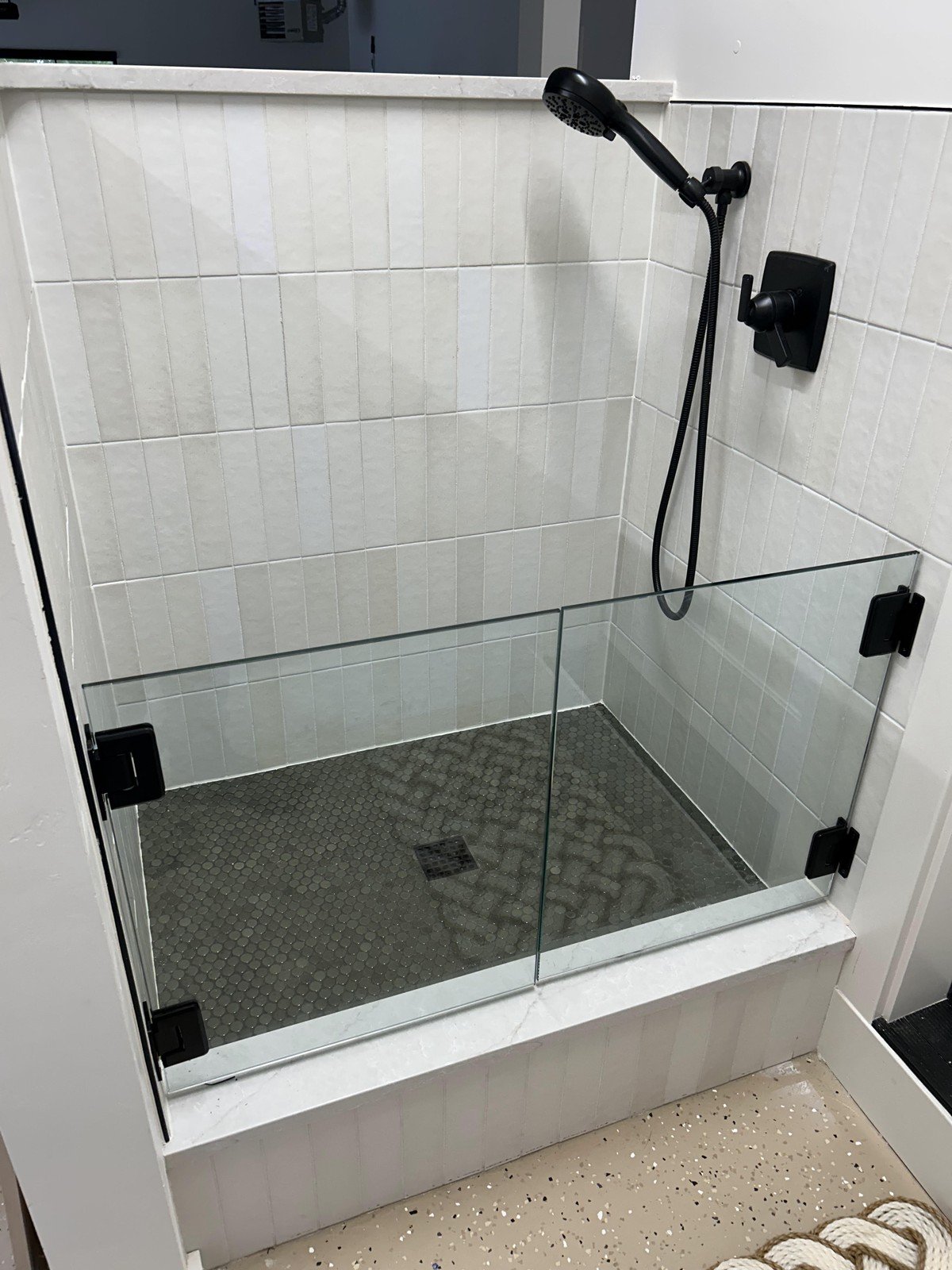 Raised shower-style dog wash enclosure with low clear glass panels and black hardware forming a partial splash barrier; white vertical subway tile, dark hex mosaic floor. No text visible in the image.