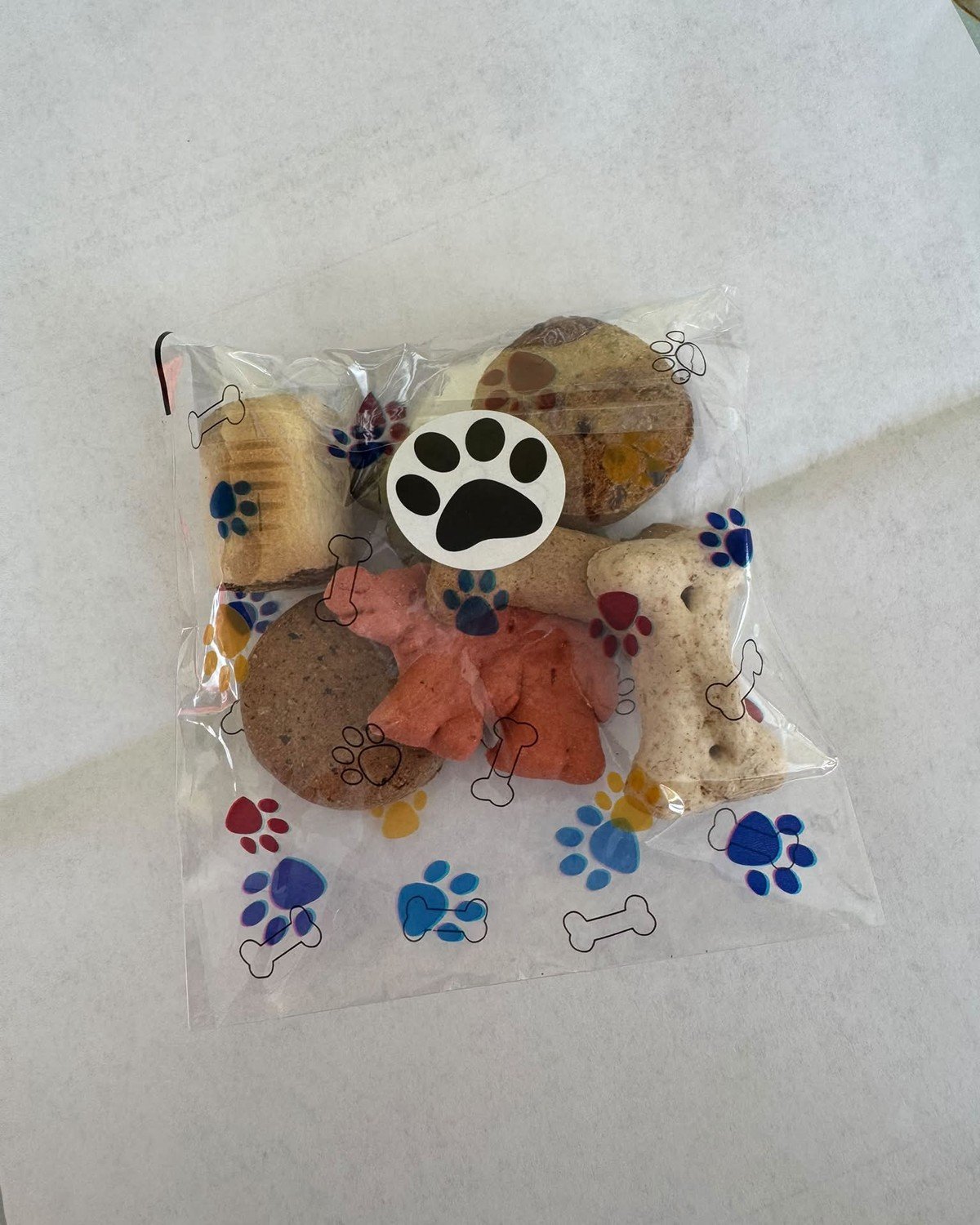 A clear favor bag filled with assorted dog biscuits, decorated with printed paw prints and sealed with a paw sticker.