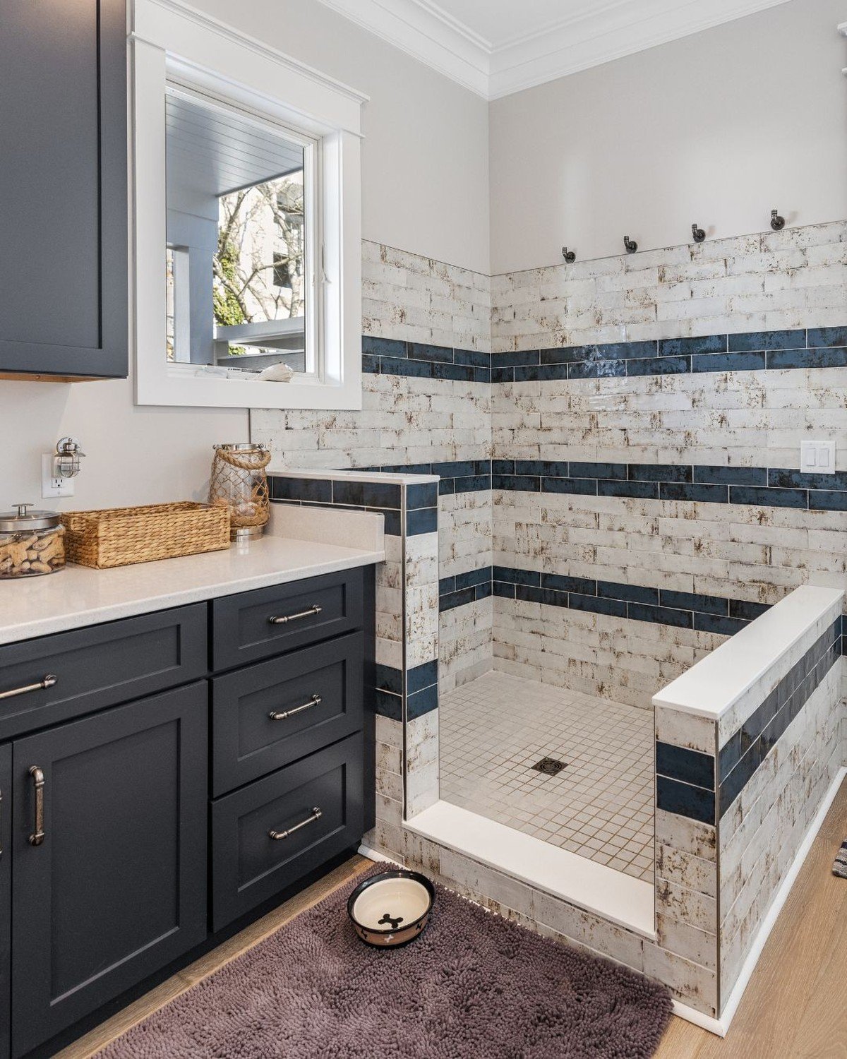 Bright mudroom with dog wash station built into dark gray shaker cabinetry run with quartz countertop; station reads as part of the cabinetry wall (copy notes adding pocket doors to close it off). No readable text.