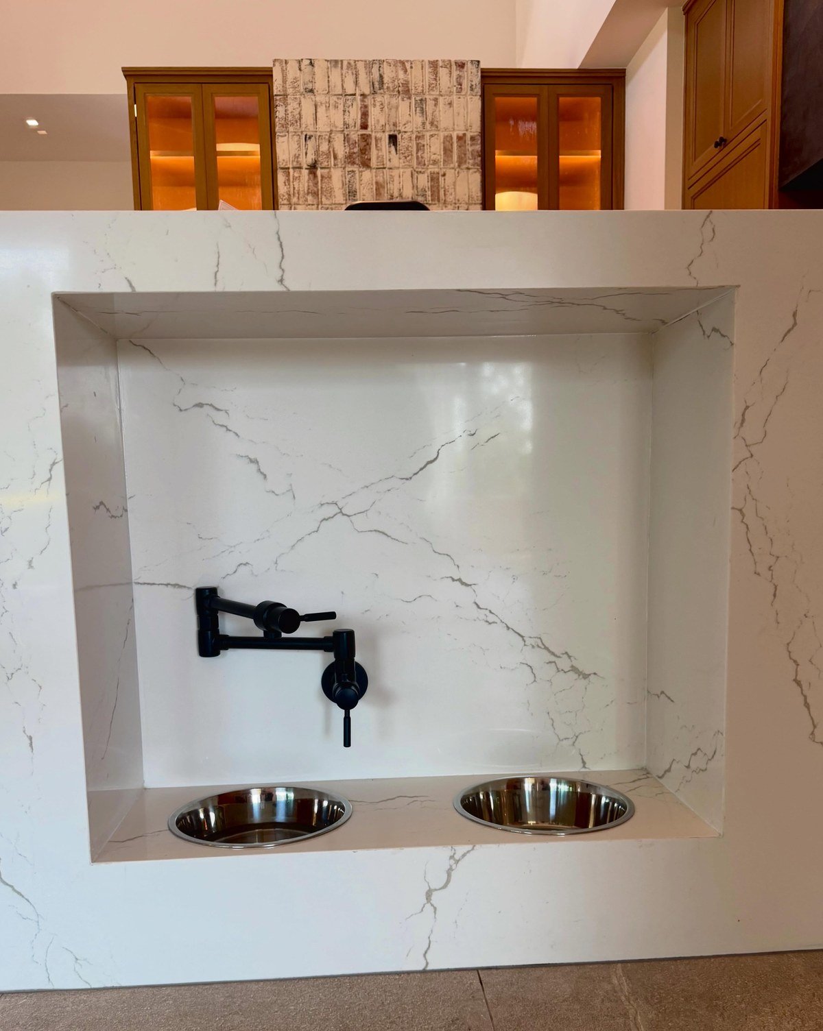 Close-up of a feeding station recessed into a white marble kitchen island, two stainless bowls set flush in the counter surface. No readable text.