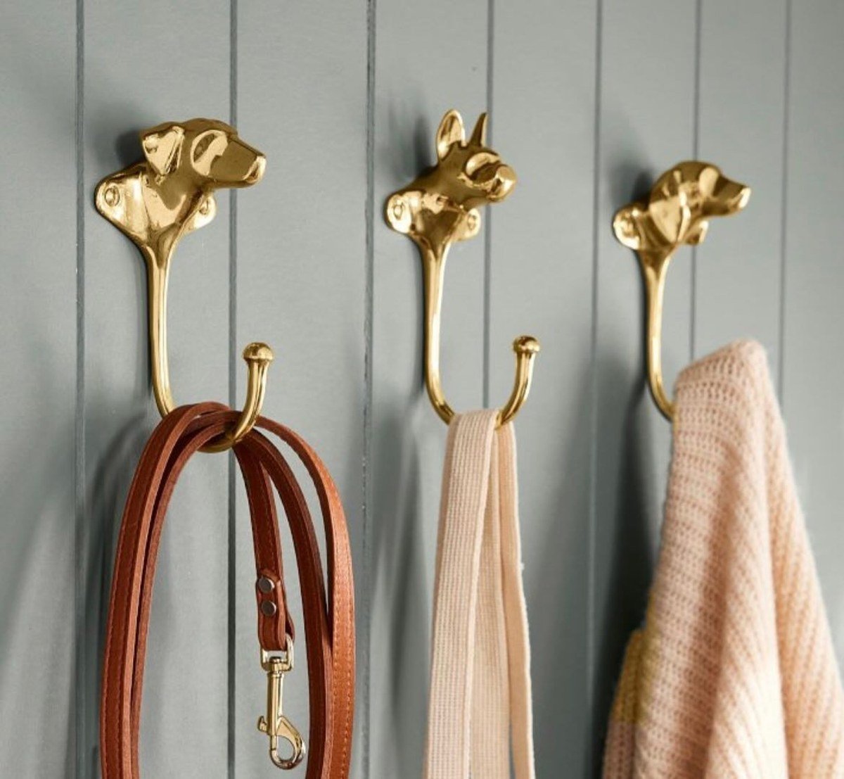 Three shiny brass wall hooks topped with sculpted dog heads mounted on a light gray paneled wall, holding a brown leather dog leash, a beige strap and a pink knitted sweater; verified no text visible in the image.