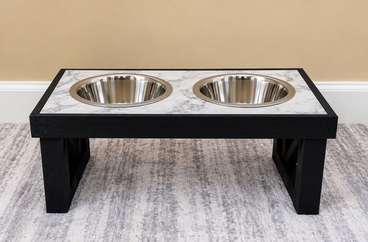 Handmade raised stand: black wooden frame with white-and-gray marble-patterned top, two stainless steel bowls, on a rug by a beige wall. No readable text.