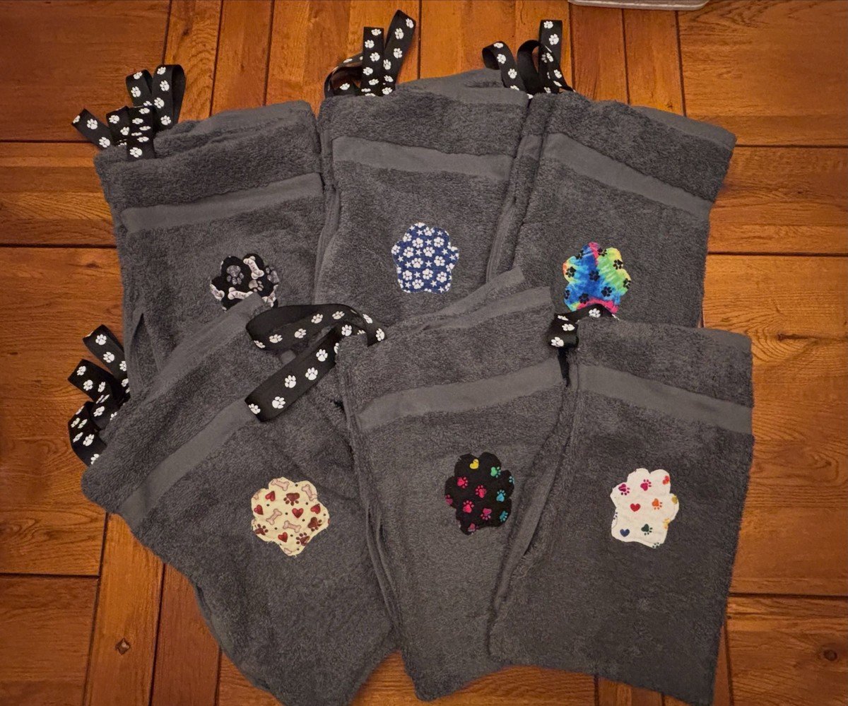 Six handmade gray paw towels with hanging loops and colorful paw patches laid out on wood floor. No readable text.