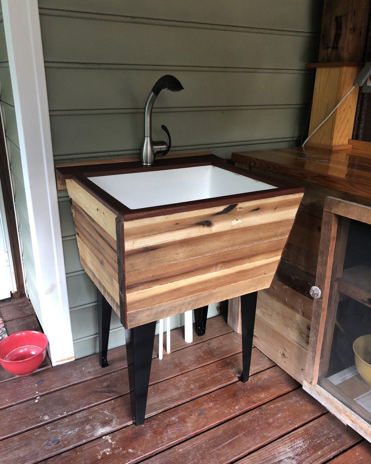 Freestanding outdoor dog washing station on a weathered wooden deck; reclaimed-wood plank basin with a stainless steel gooseneck faucet and pull-down spray head. No text visible in the image.