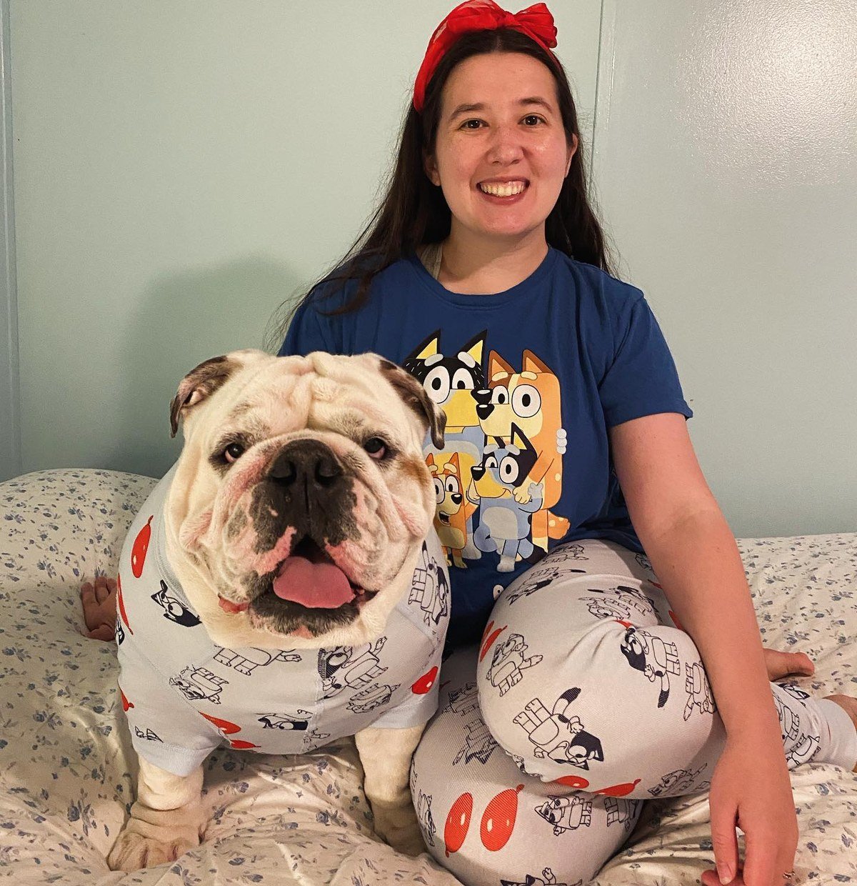 Bulldog wearing cartoon pajamas sitting on a bed with its owner in matching pajamas