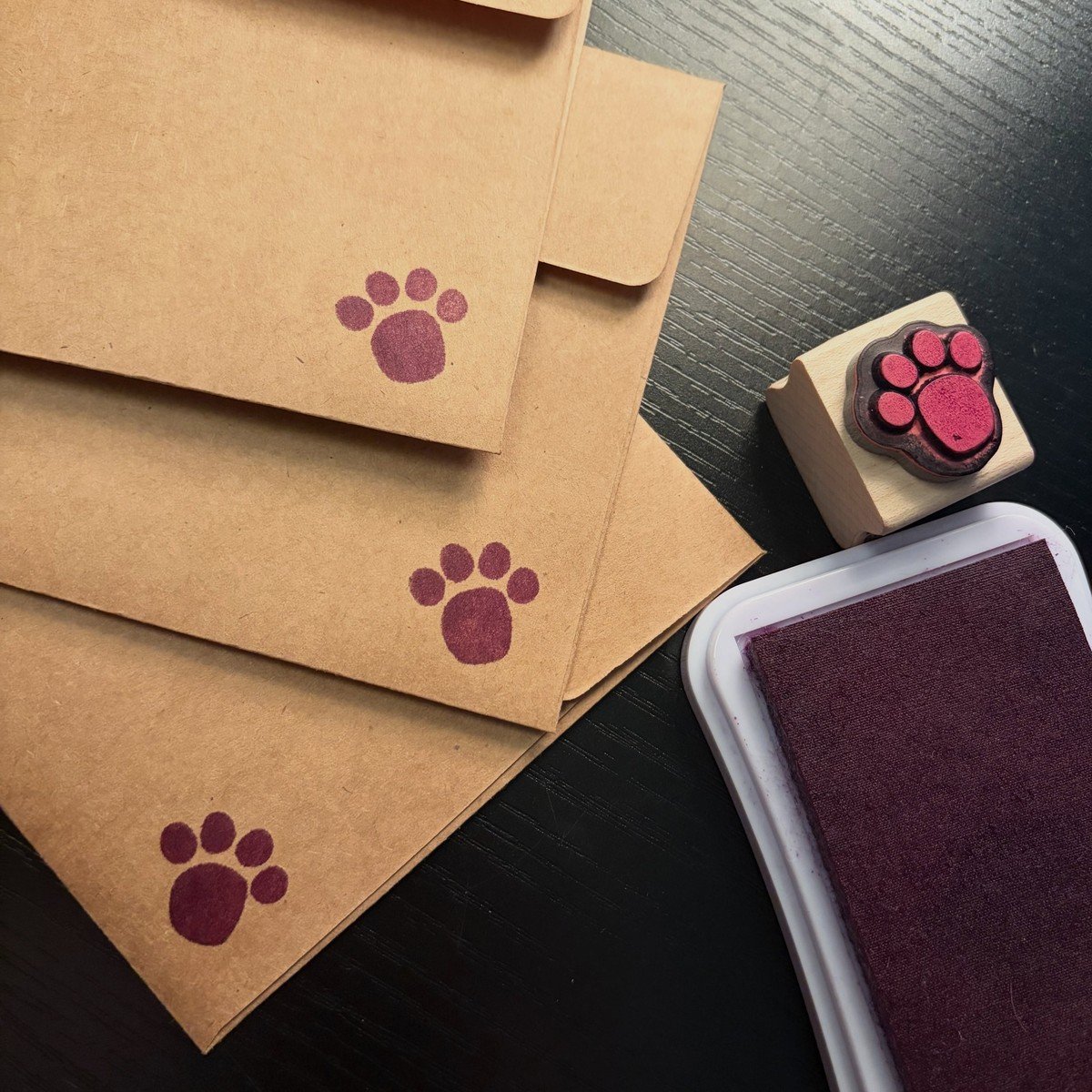 Three kraft envelopes stamped with burgundy paw prints beside a matching paw-print rubber stamp and ink pad.