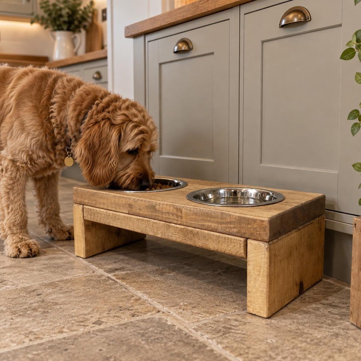 Curly-haired dog eating kibble from a stainless bowl set in a handmade raised wooden feeder on a stone-tiled kitchen floor. No readable text.