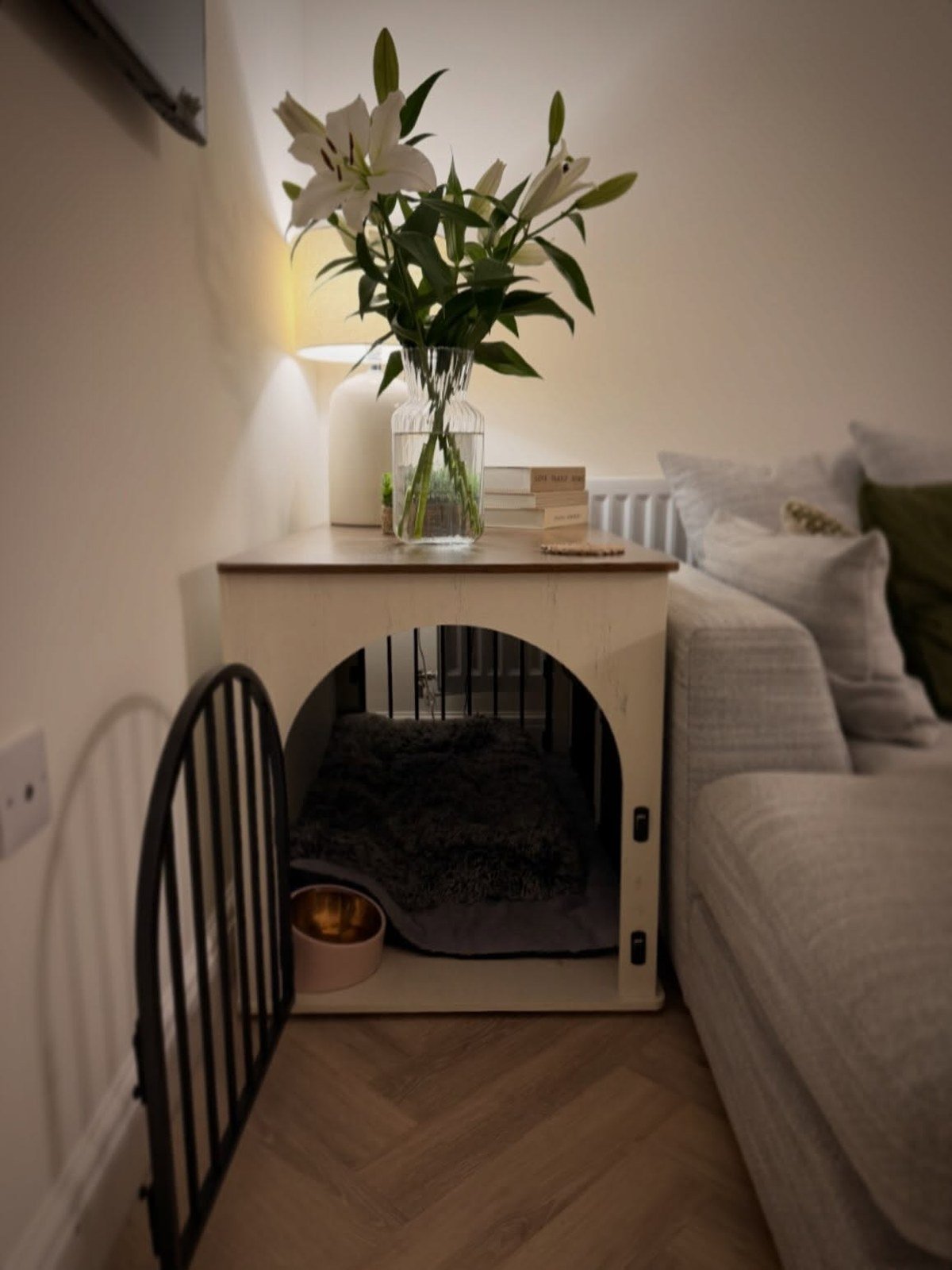 Custom wooden dog crate doubling as a side table, styled with a vase, lamp, and books beside the sofa.