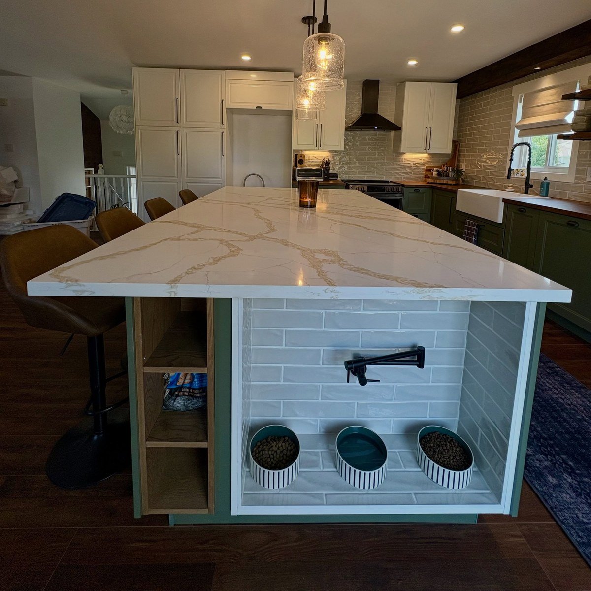 Tiled alcove under a kitchen island overhang with a low wall-mounted pet faucet and three striped ceramic bowls below. No readable text.