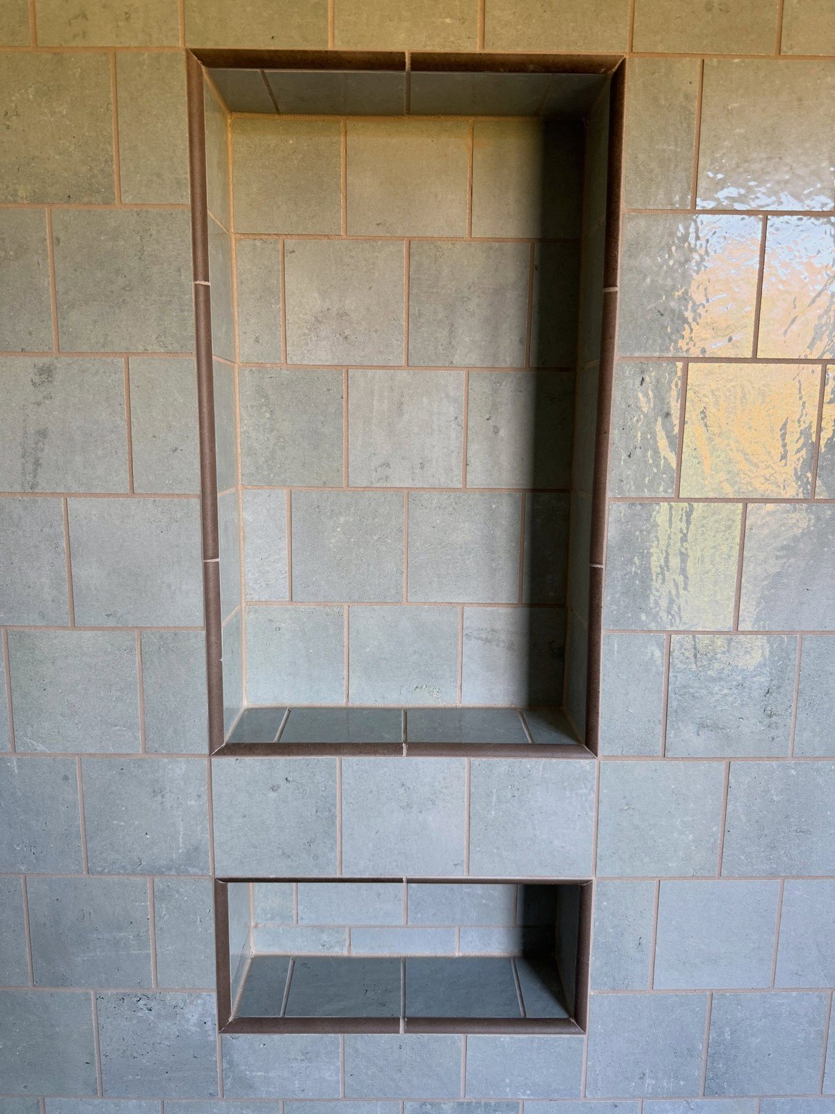 Close-up of a tiled shower wall with two built-in recessed niches in muted sage-green and gray tile; empty shelves, no bottles. No text visible in the image.