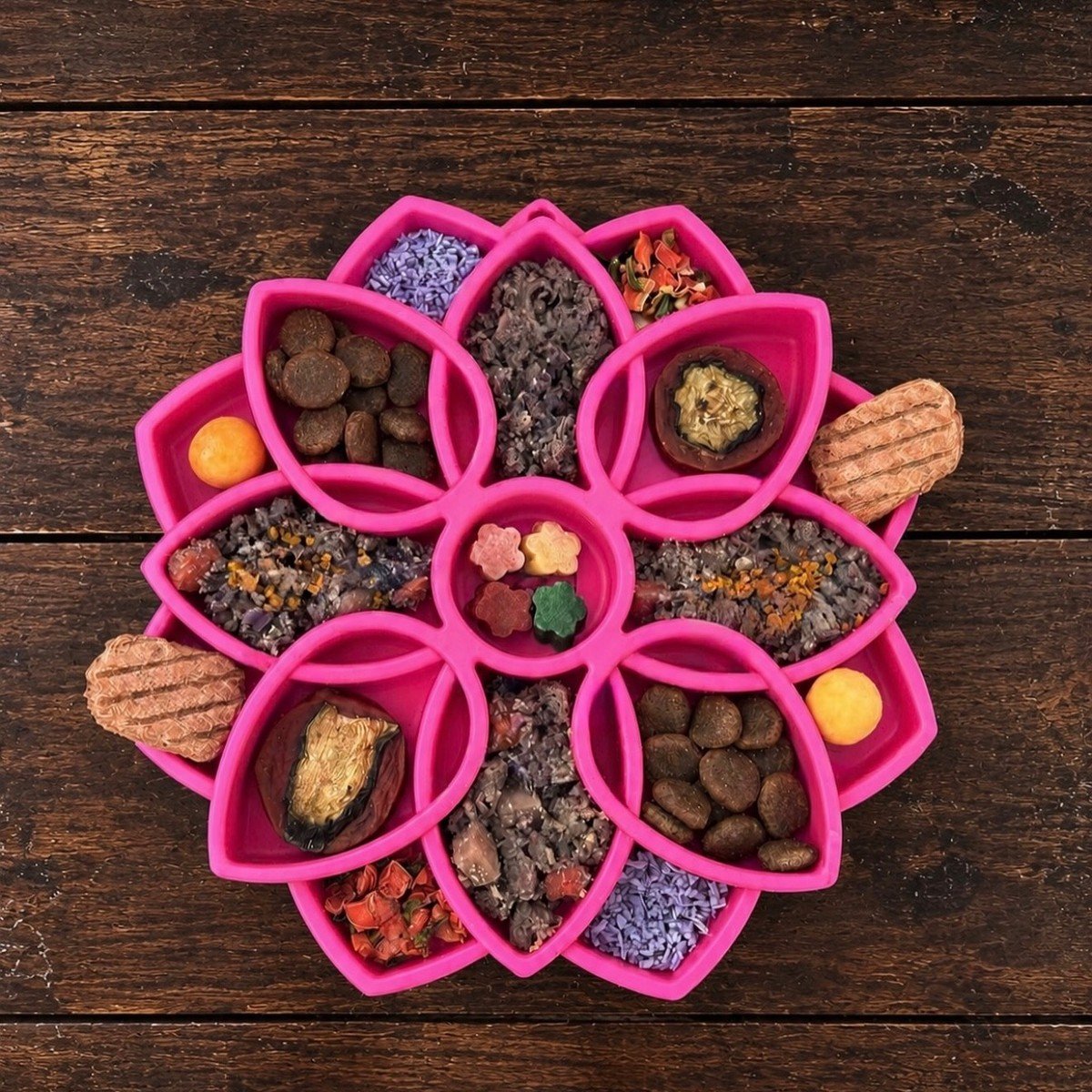 A pink flower-shaped silicone tray holds a colorful mandala-style spread of dog treats, kibble, and wet food.